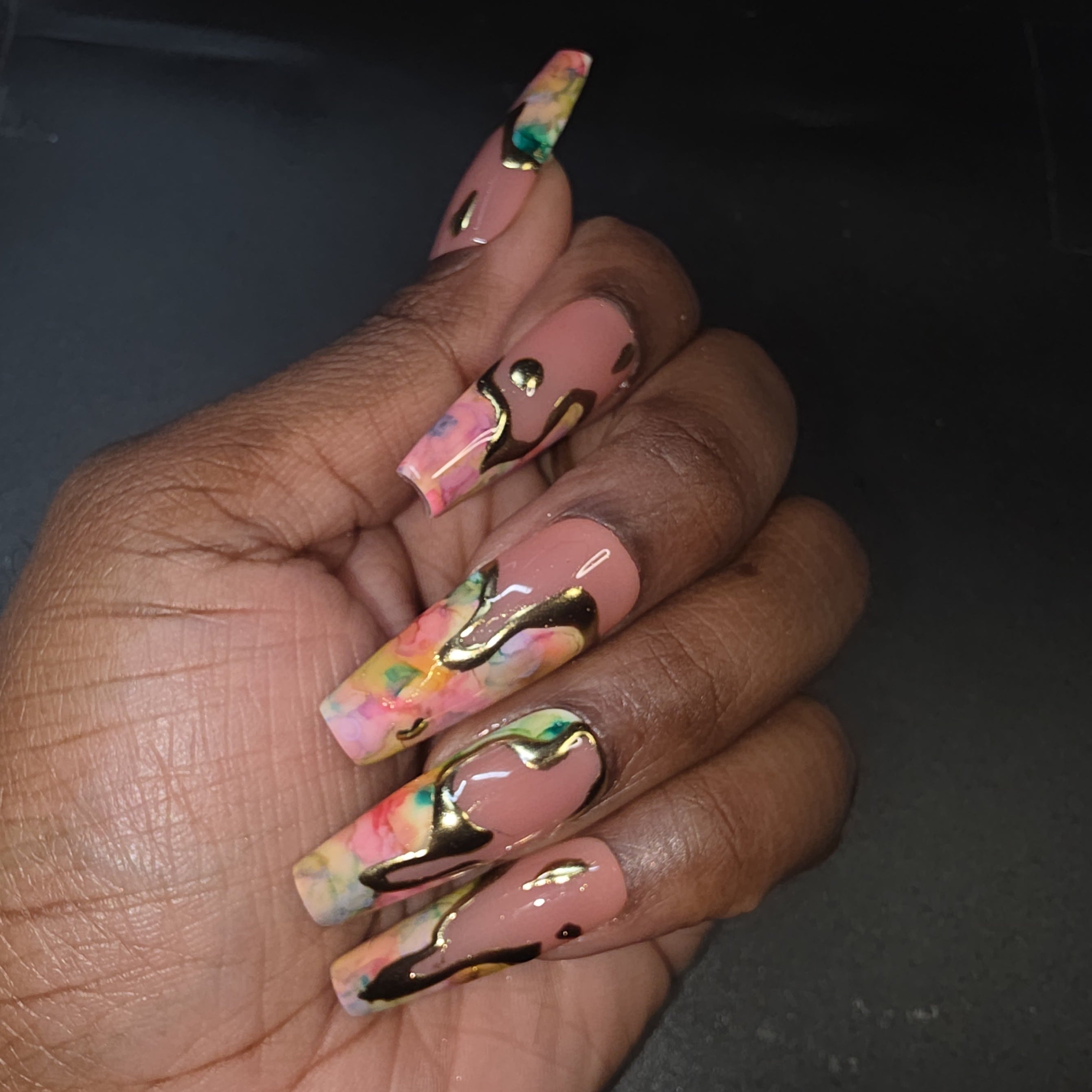 Hand with colorful acrylic nails featuring gold accents on a dark background